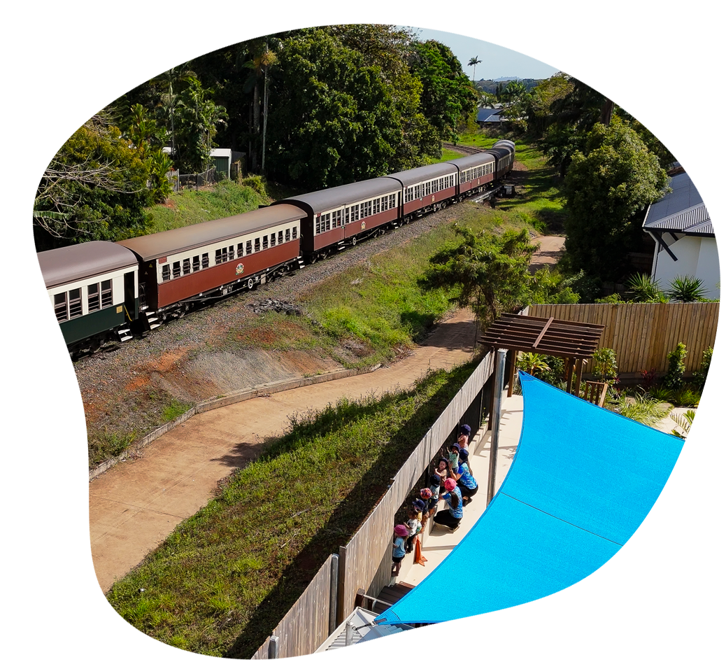 Train traveling along tracks near green trees and a shaded seating area.