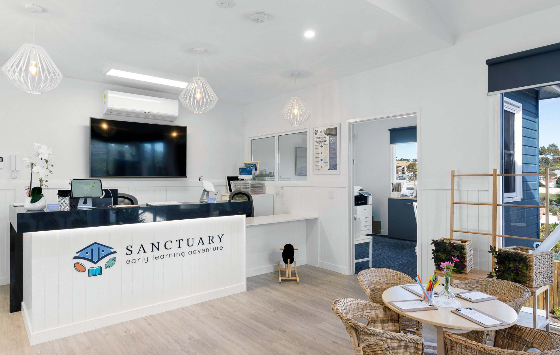 Reception area with desk, logo, and waiting area with tables and chairs. Bright, airy space with a view.