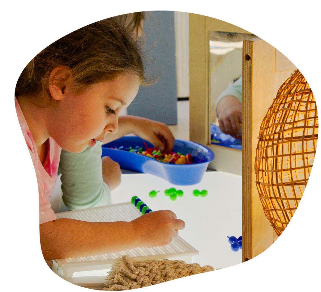 Girl writing on a light table, playing with colorful beads; a mirror reflects her activity.