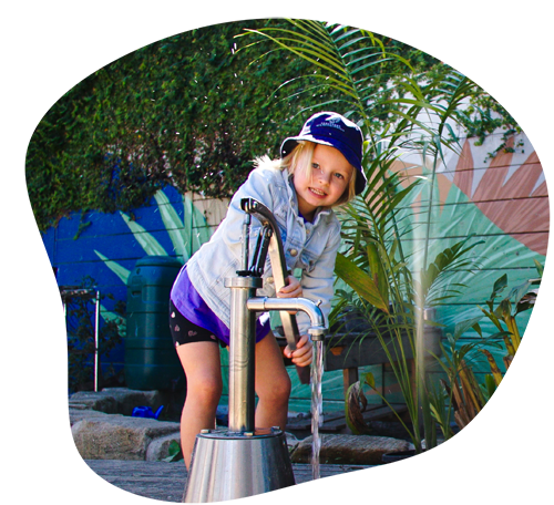 Child playing with a water pump, smiling, outside with greenery.