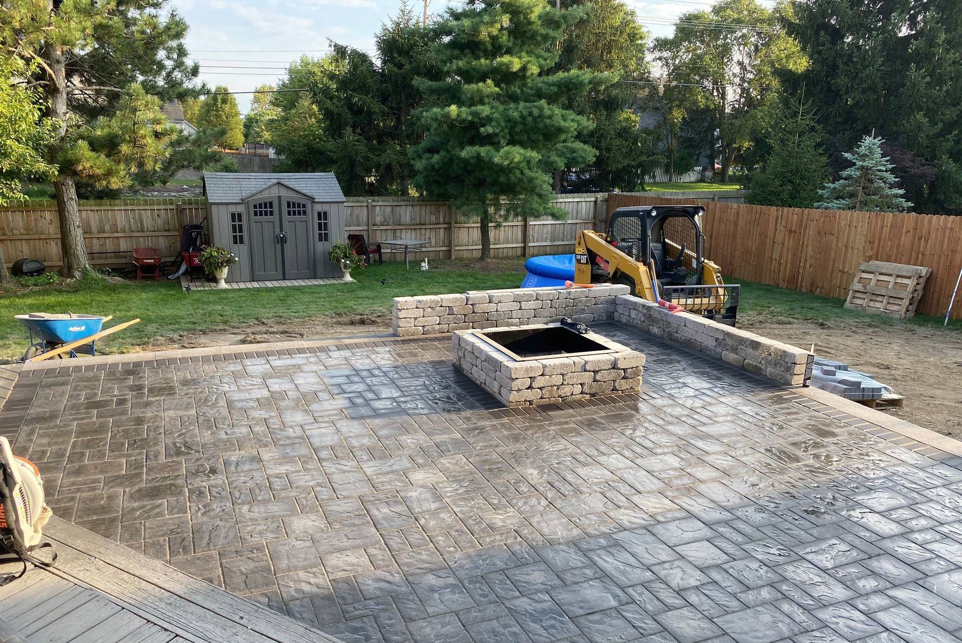 Backyard patio with fire pit under construction; excavator in the background, trees, and shed in the yard.