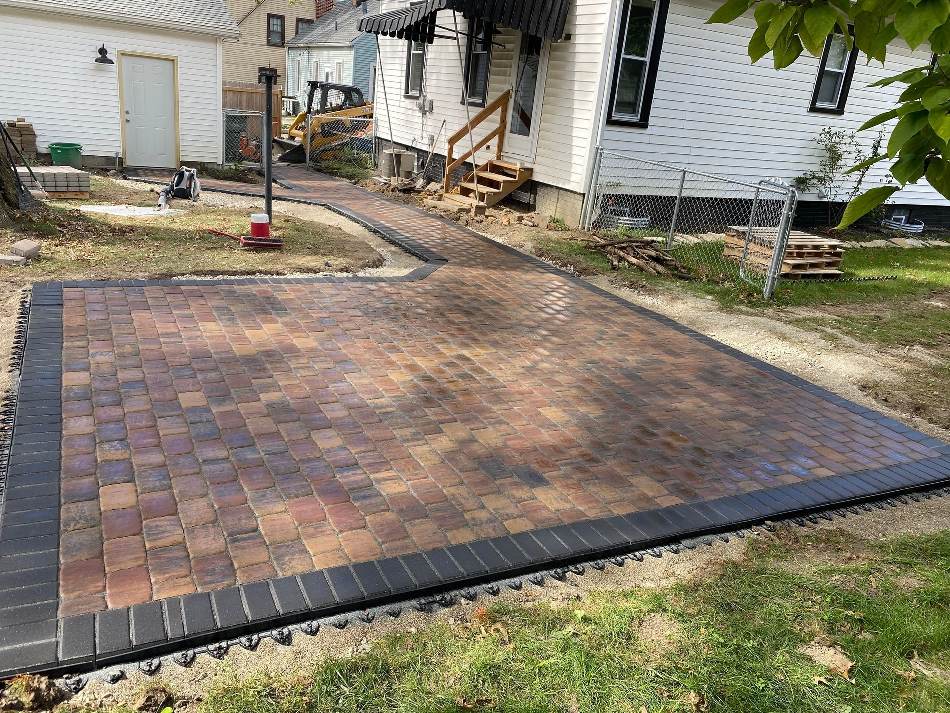 Brick patio with dark border, leading to a sidewalk in front of a white house.