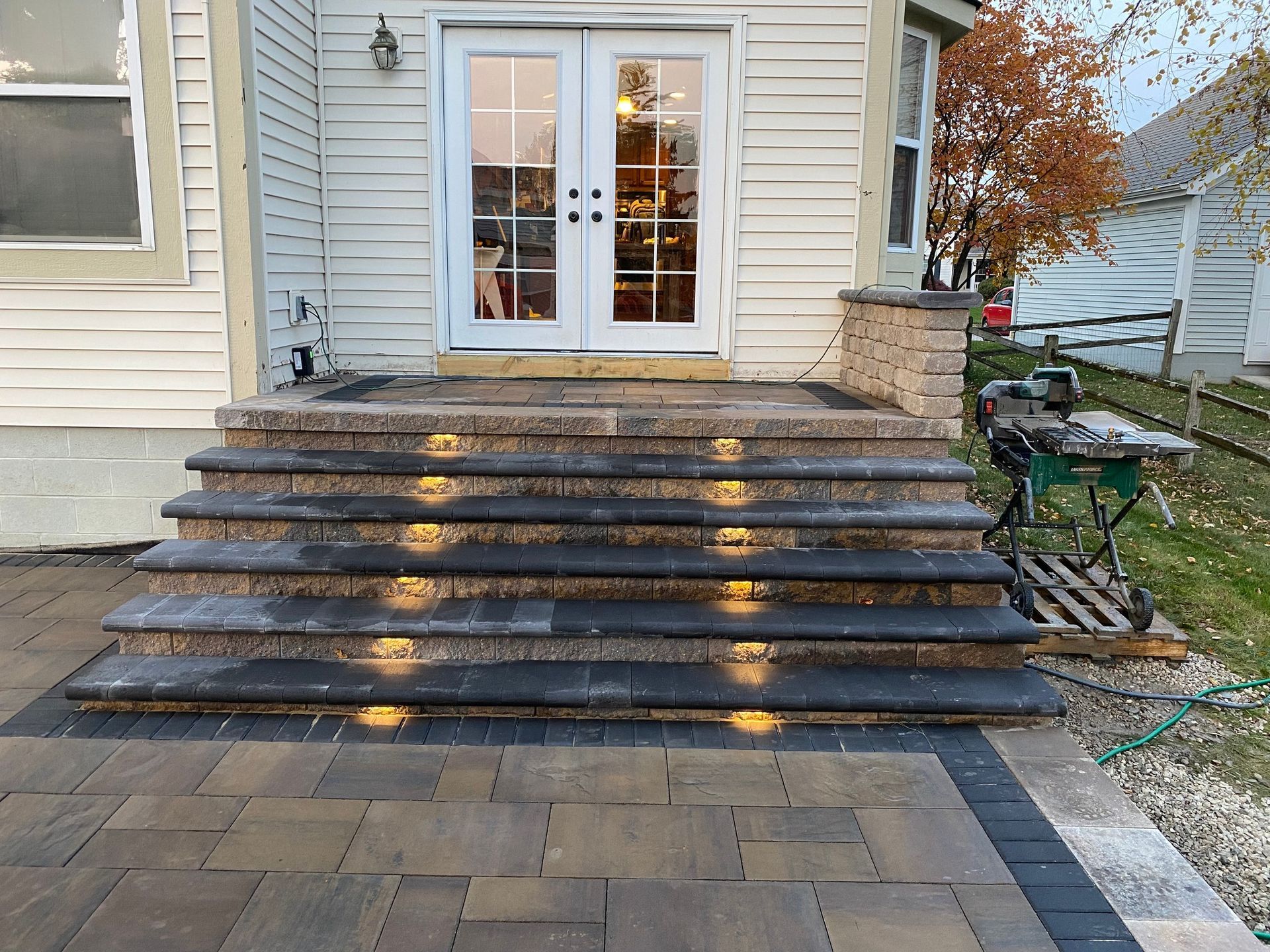 Stone steps with built-in lights lead to a house with double doors, set on a paved patio, with a lawn in the background.