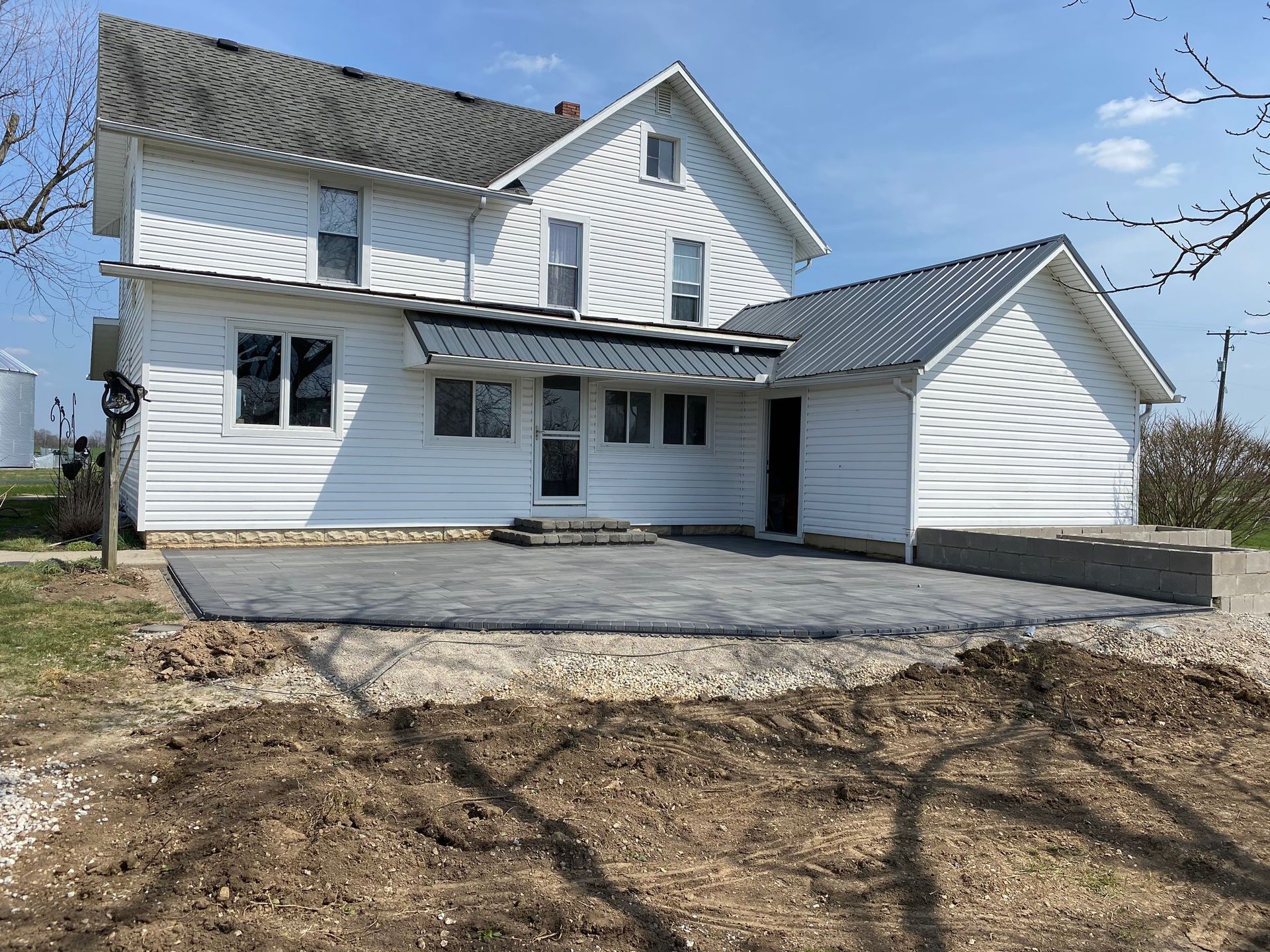 White farmhouse with new patio and detached garage on a sunny day. Dirt and gravel in foreground.