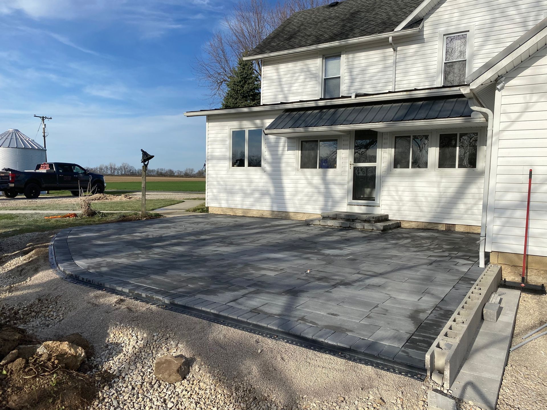 Newly constructed gray paver patio next to a white farmhouse.