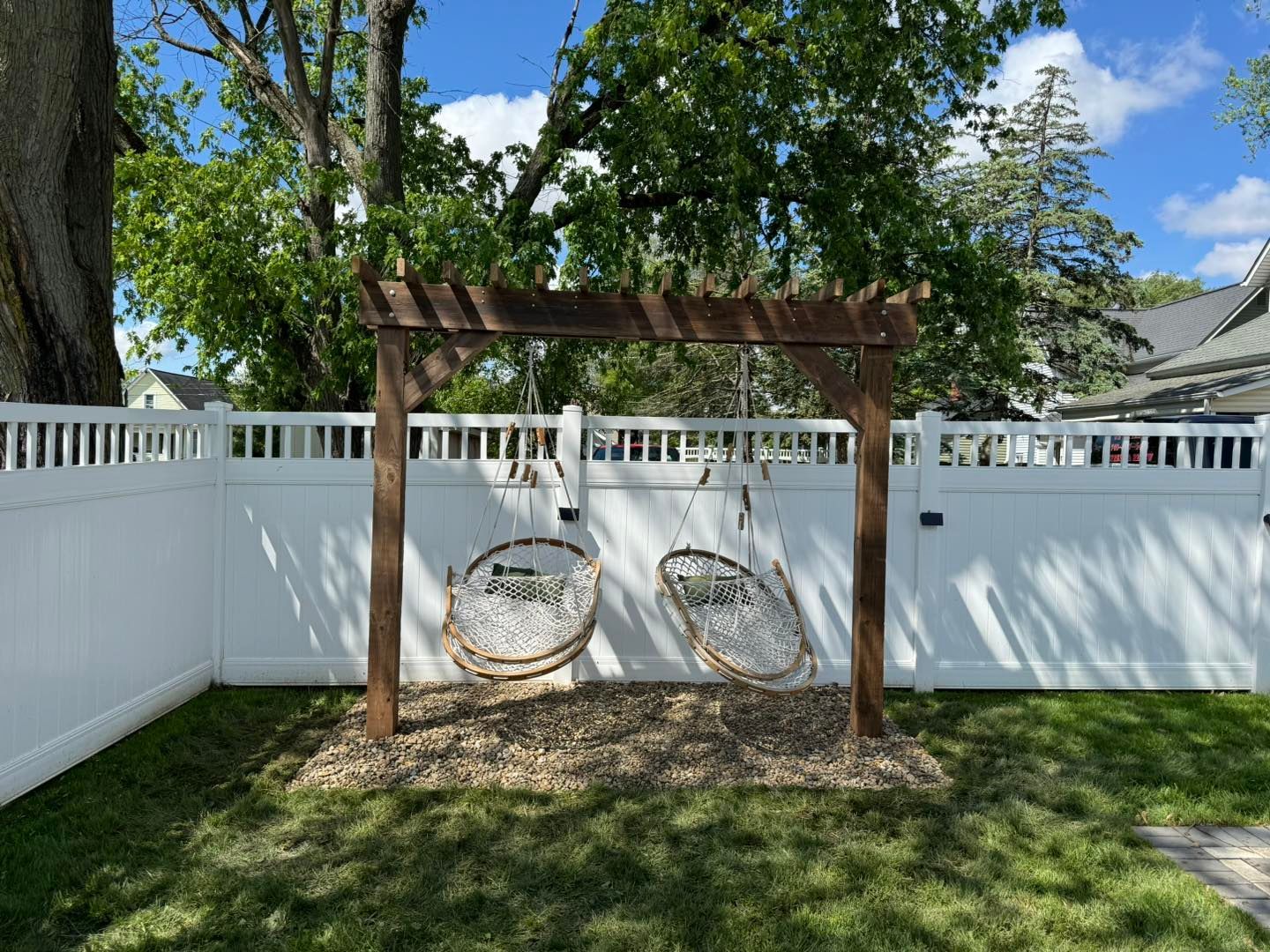 Two hanging chairs under a brown wooden pergola in a backyard with a white fence.