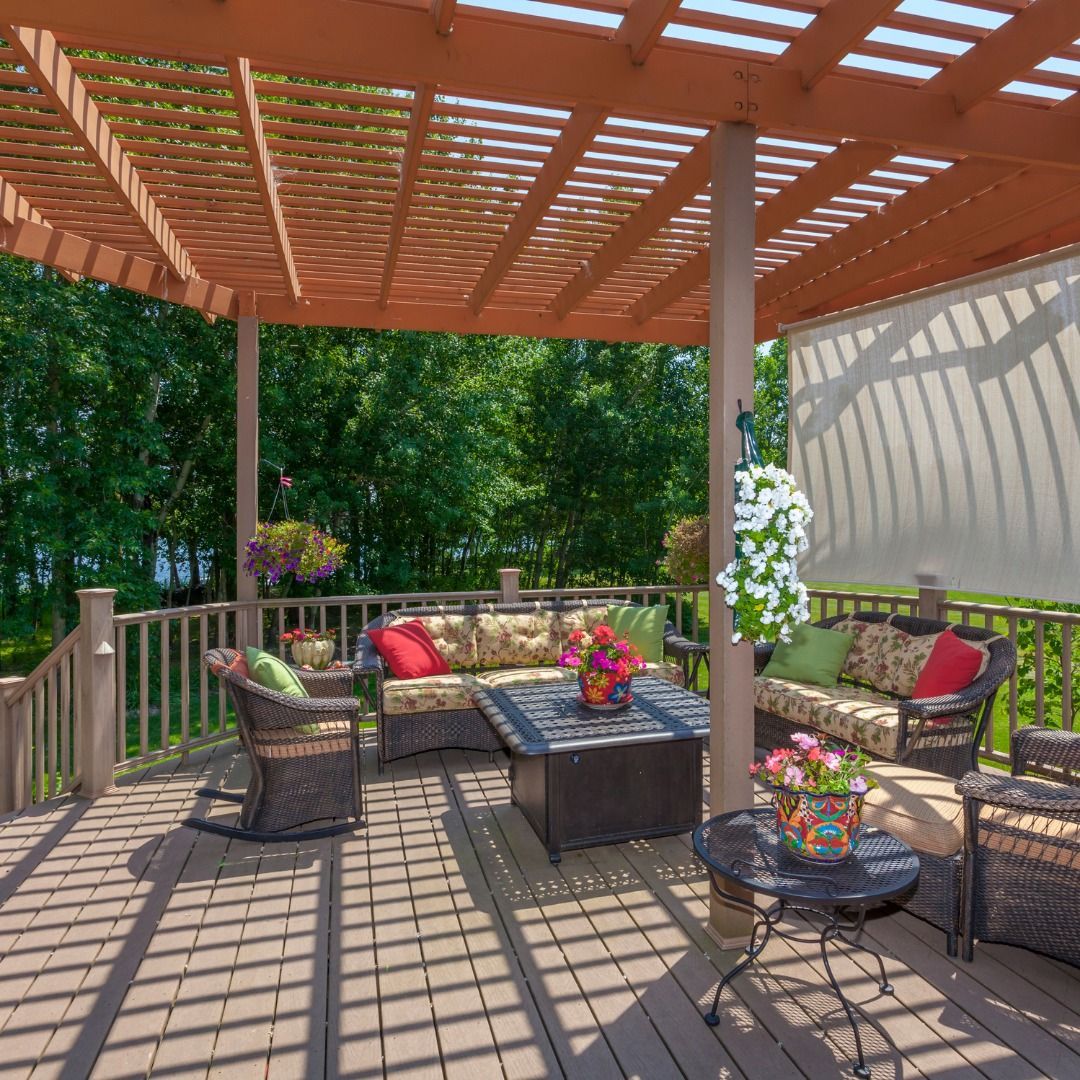 Outdoor deck with lounge seating, pergola, and hanging flowers; sunny day.