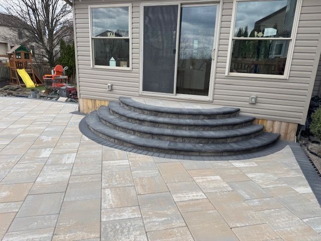 Stone patio with curved steps leading to a sliding glass door, beige siding, and surrounding landscaping.