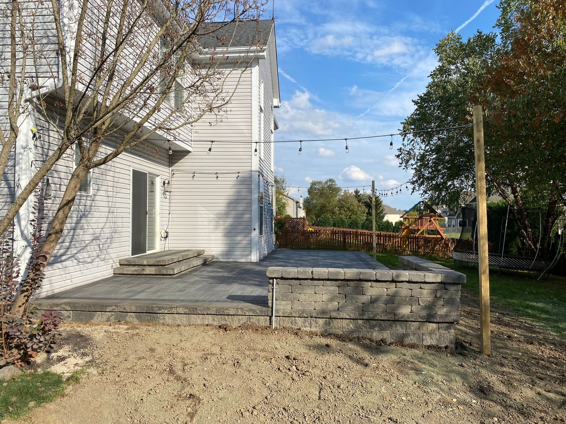 Backyard patio with gray pavers and retaining wall, attached to a white house with string lights.
