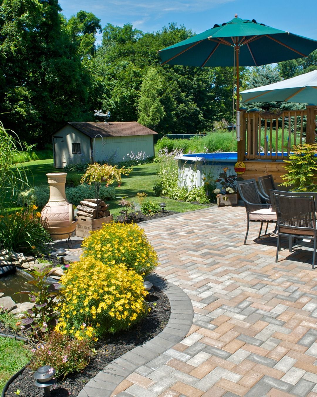 Brick patio with yellow flowers, water feature, and pool in a backyard.