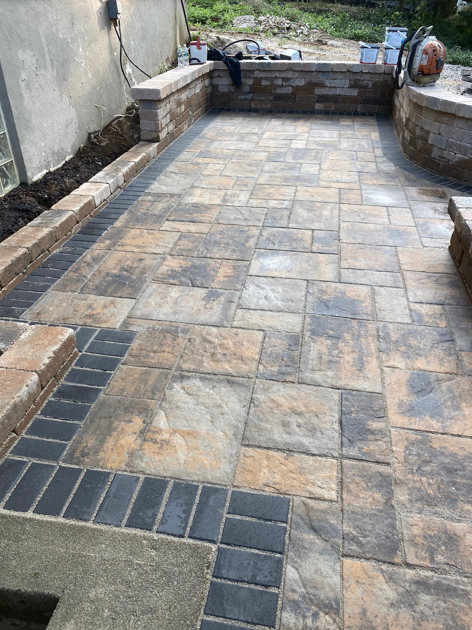 Stone patio under construction with tan and gray pavers. Dark gray border, stone retaining walls.