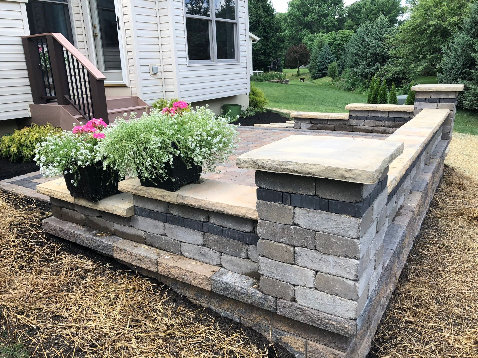 Stone retaining wall with flowers on a patio in front of a house.