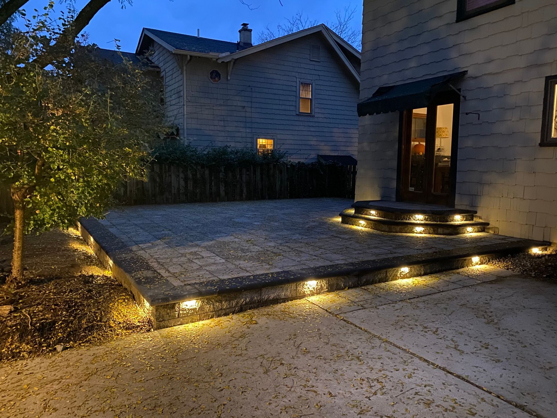Evening view of a brick patio and steps illuminated by warm lights, next to a house with a dark green awning.