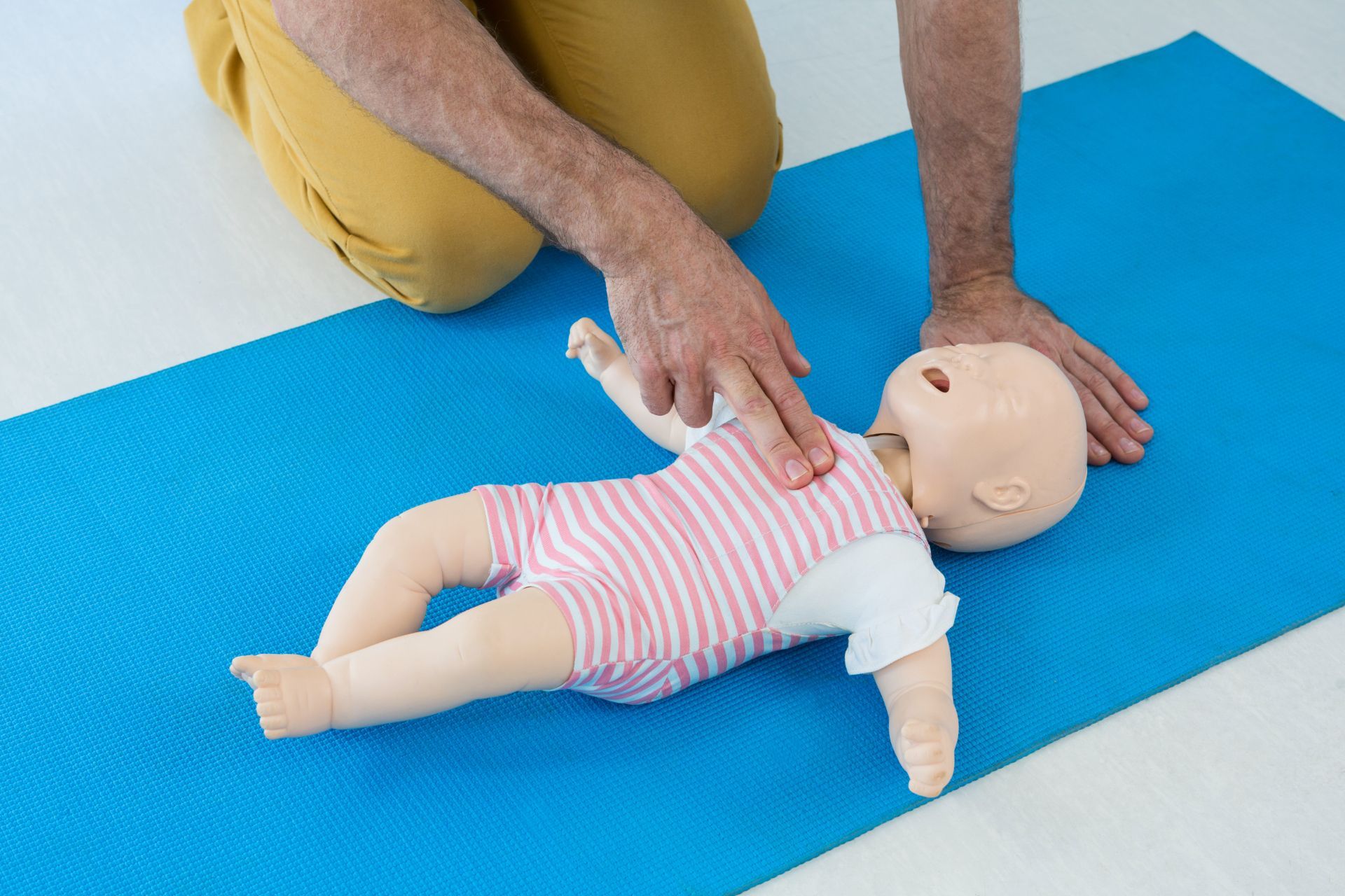 A man is kneeling down next to a baby mannequin for CPR on a blue mat.