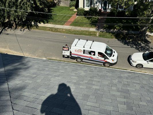 White van and car parked on a street next to a house with a green lawn.