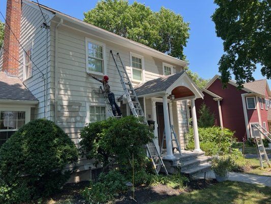 Person on ladders painting the exterior of a two-story beige house on a sunny day.