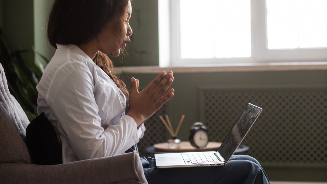 Woman gestures with hands during a video call on laptop, sitting in chair near window.
