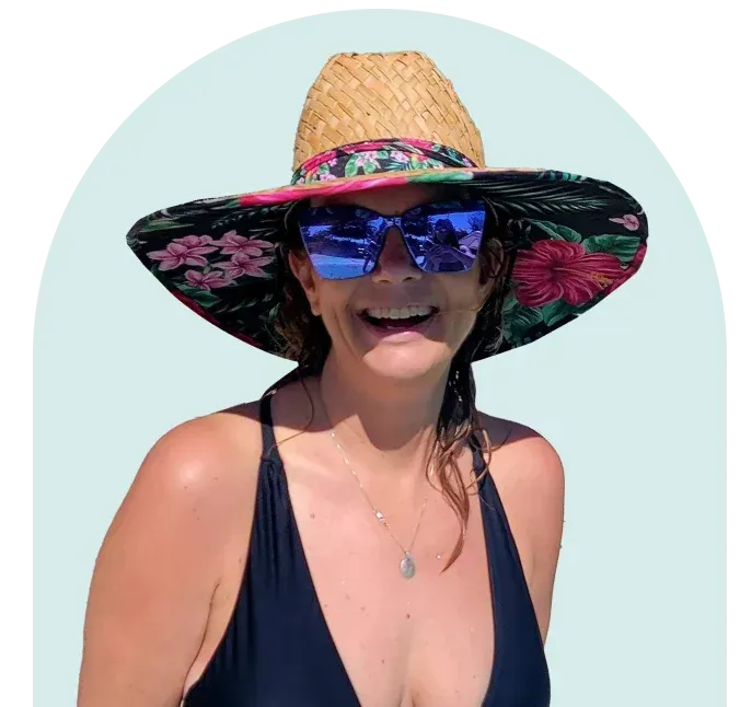 Woman wearing a wide-brimmed floral hat and blue sunglasses, smiling. She's in a swimsuit, likely outdoors near water.