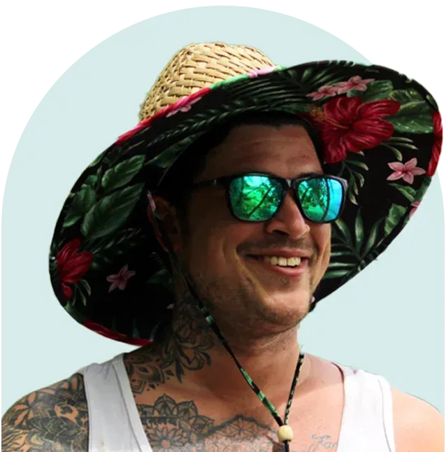 Man wearing floral sun hat and sunglasses, smiling, with tattoos visible on his arms and neck.