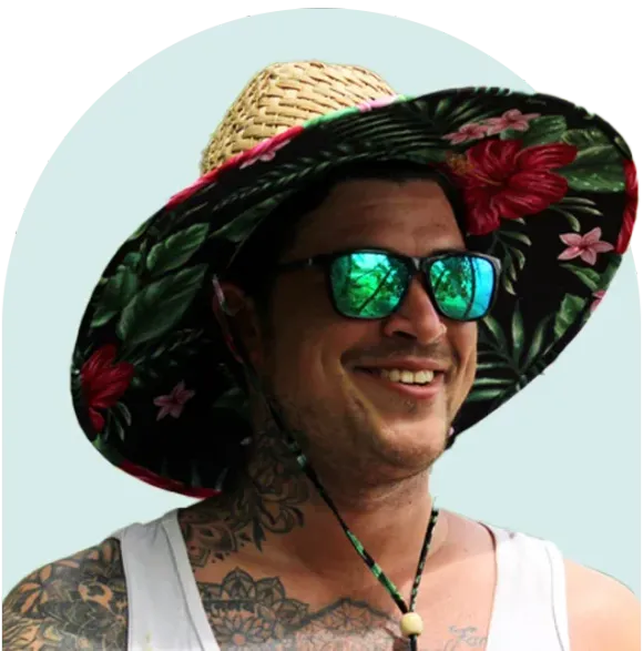 Man wearing floral sun hat and sunglasses, smiling, with tattoos visible on his arms and neck.