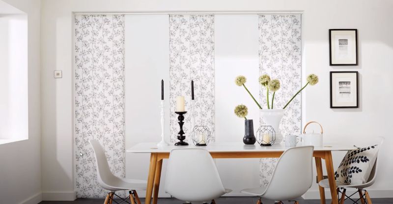 A clean, white dining room has layered window treatments with patterned drapes. 