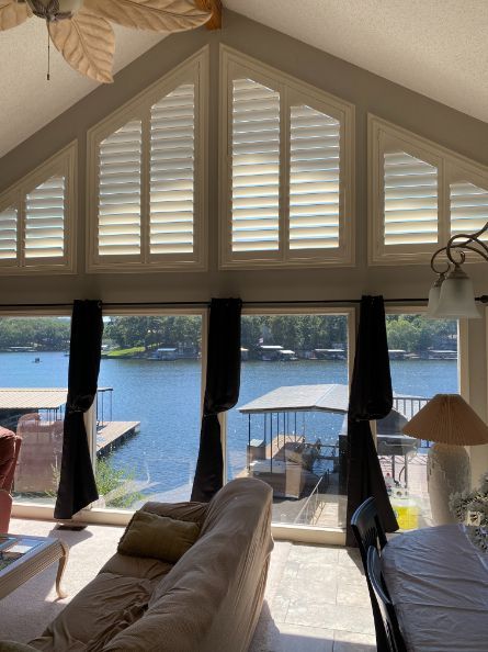 A living room has large windows that look out over a lake with boats, and white shutters are on the 