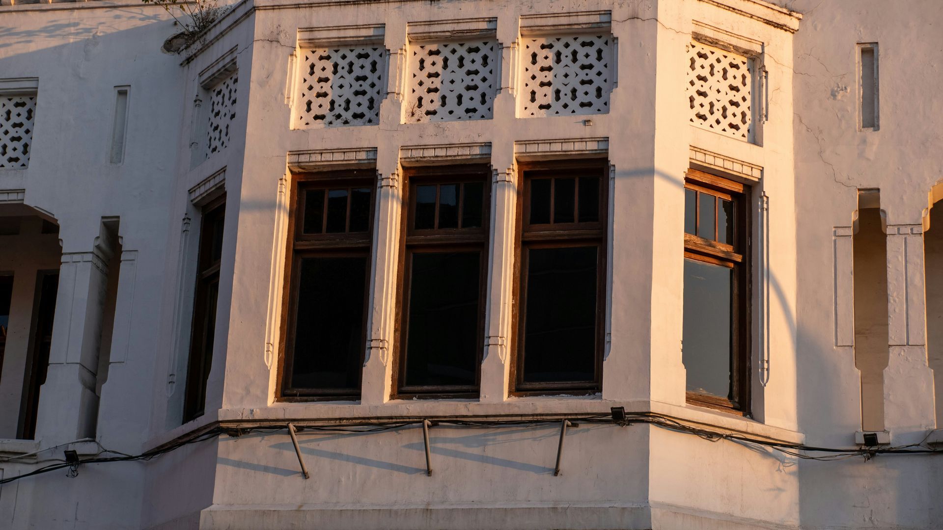 White building facade with dark windows and decorative top panels, lit by golden sunlight and window treatments