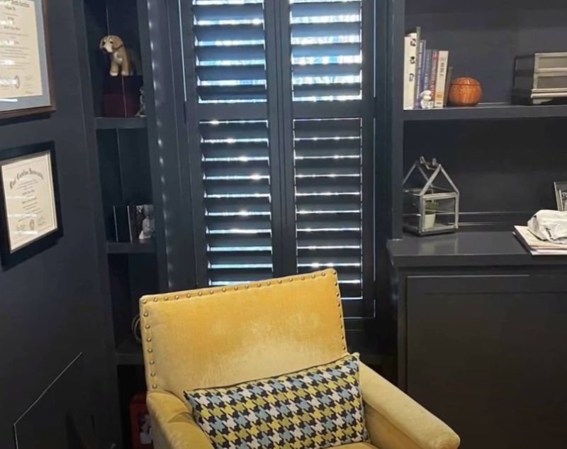 A chair with a yellow pillow is sitting in a room with shutters on the windows.