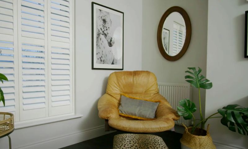 A living room with a chair , mirror , shutters and a picture on the wall.