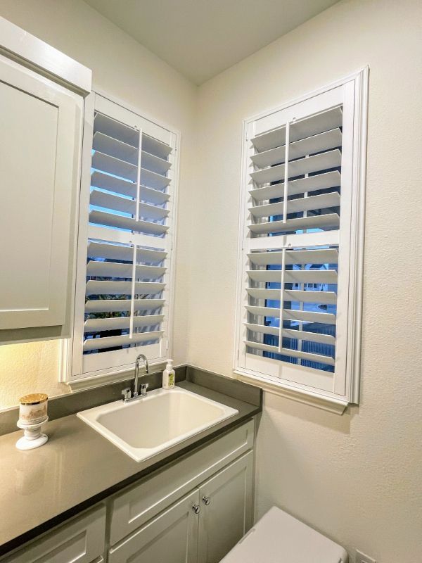 A bathroom with a sink , toilet and two windows with shutters.