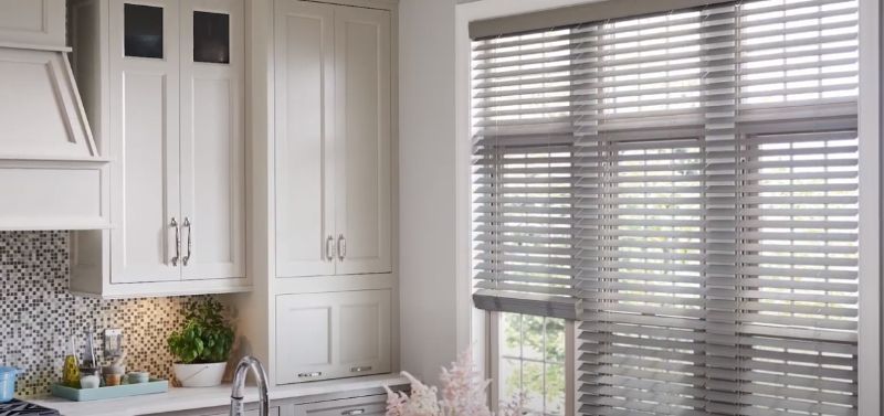 A kitchen with white cabinets , a sink , and a window with blinds.