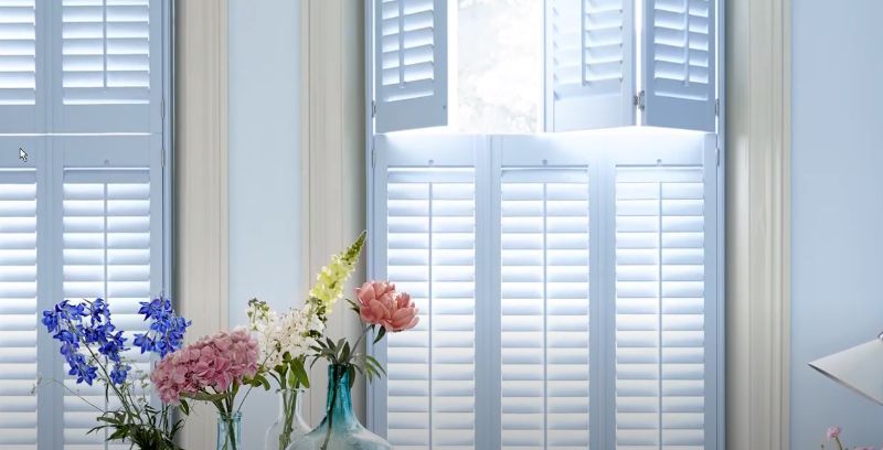 A vase of flowers sits on a table in front of a window with white shutters.