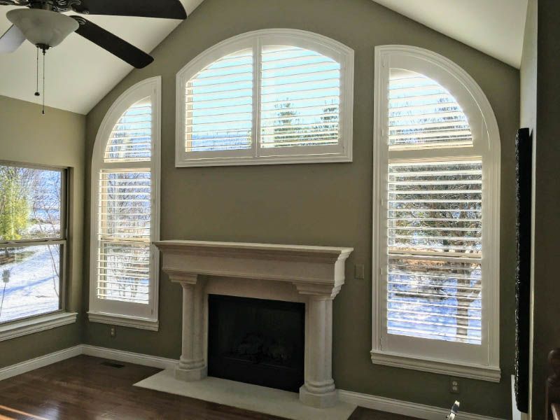 Large, arched windows with plantation shutters are designed around a fireplace.