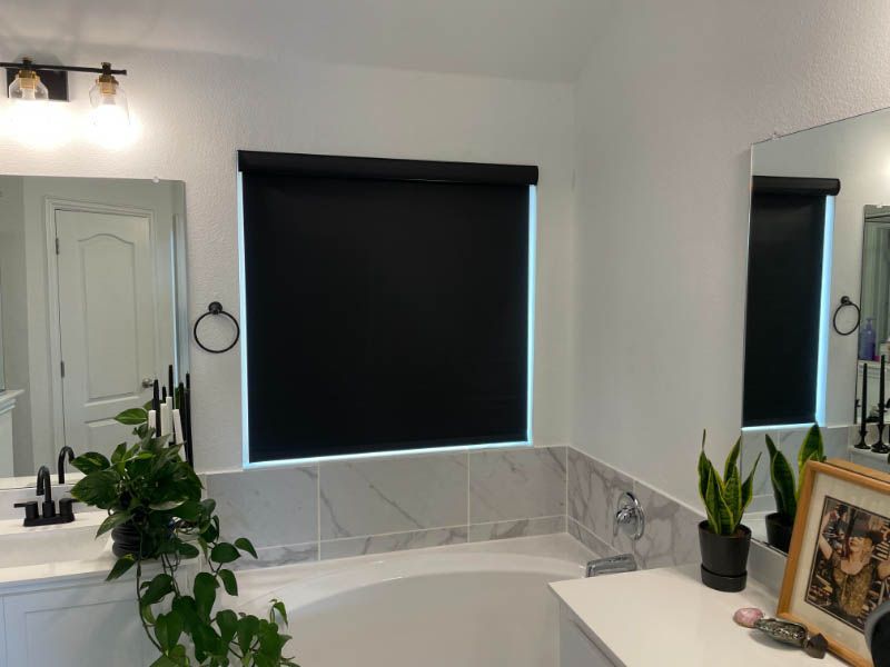 Black roller shades cover a window over a bath tube in an elegant bathroom.