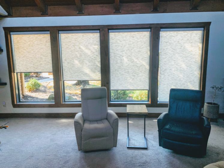 A den with two chairs has a wall of windows with roller shades. 