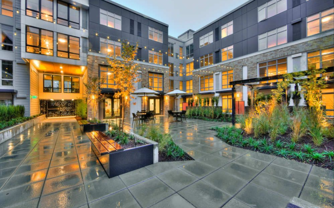 Courtyard of modern apartment complex with outdoor seating, plants, and string lights.