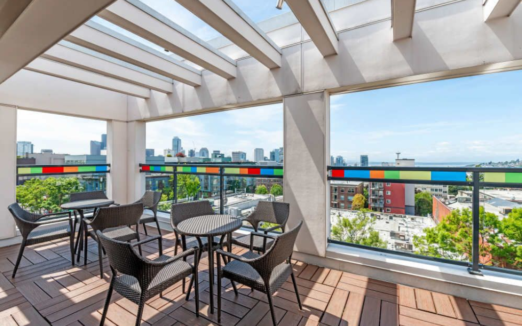 Rooftop patio with city view. Black chairs and tables under a pergola, colorful glass railing, blue sky.