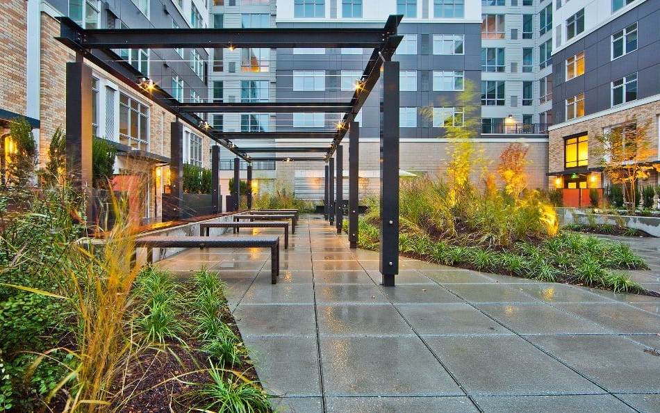 Outdoor patio with metal pergola, tables, and landscaping in an urban setting.