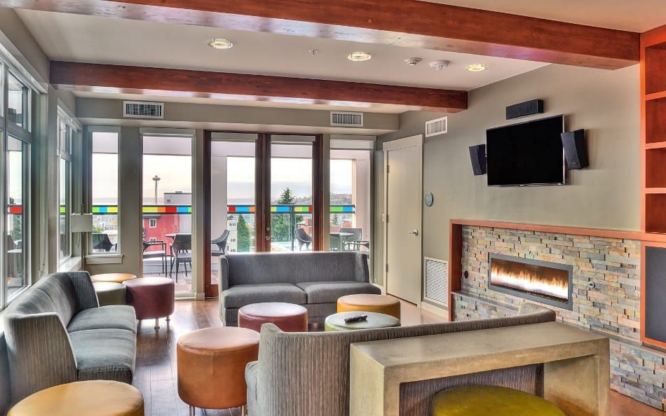 Lounge area with gray sofas, brown ottomans, fireplace, TV, wooden beams, and large windows with a view.