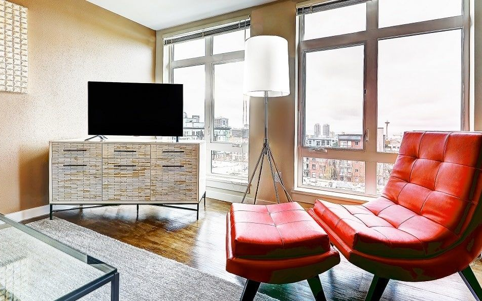 Living room with red chair and ottoman, TV on white cabinet, and floor lamp by a large window.