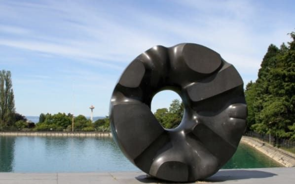 Large dark sculpture in donut shape sits on a wall overlooking a body of water and trees under a blue sky.
