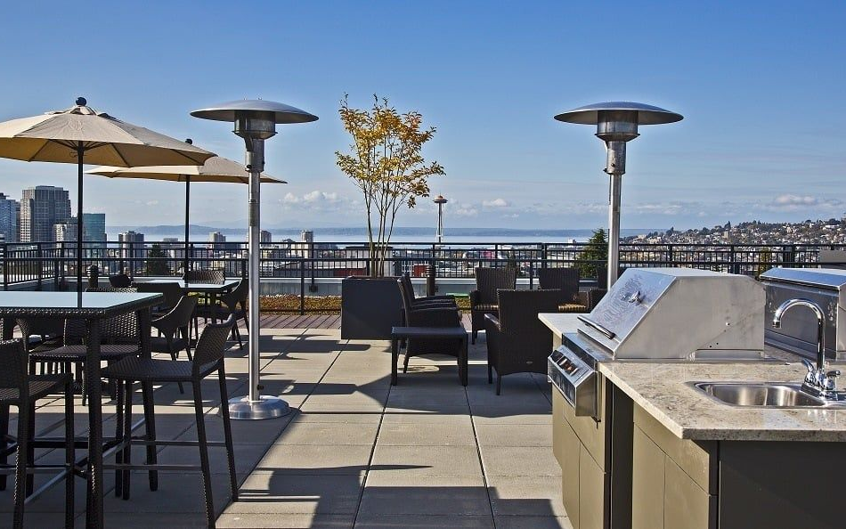 Rooftop deck with grilling station, seating, umbrellas, and heaters overlooking a city and water on a sunny day.