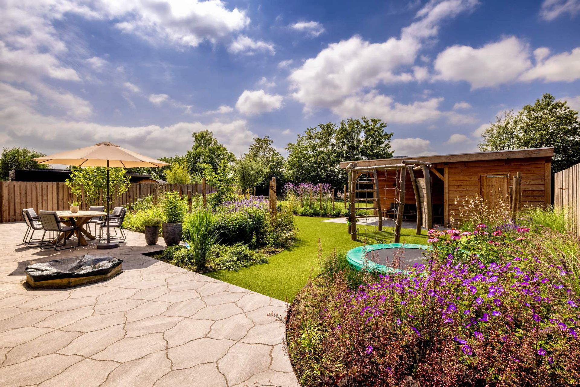 Een zonnige achtertuin met een stenen terras, tuin, houten schuur, speeltoestel en eethoek onder een parasol.