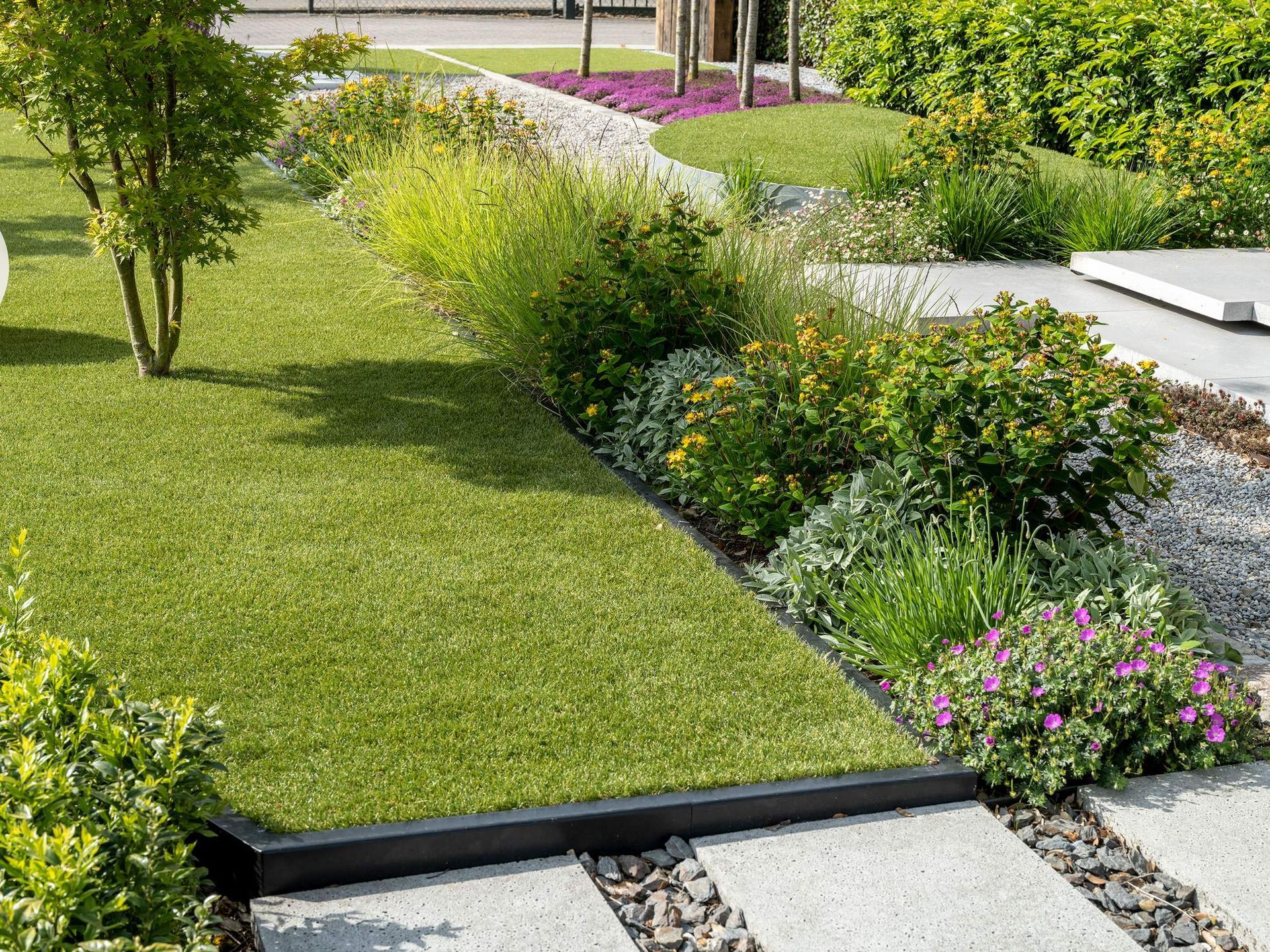 Weelderige, groene aangelegde tuin met kunstgras, verschillende planten, grind en betonnen stapstenen.