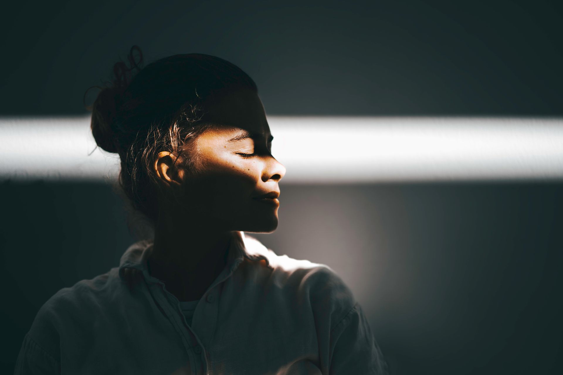 Woman in partial shadow, eyes closed, looking serene. Soft light on face and shoulder.