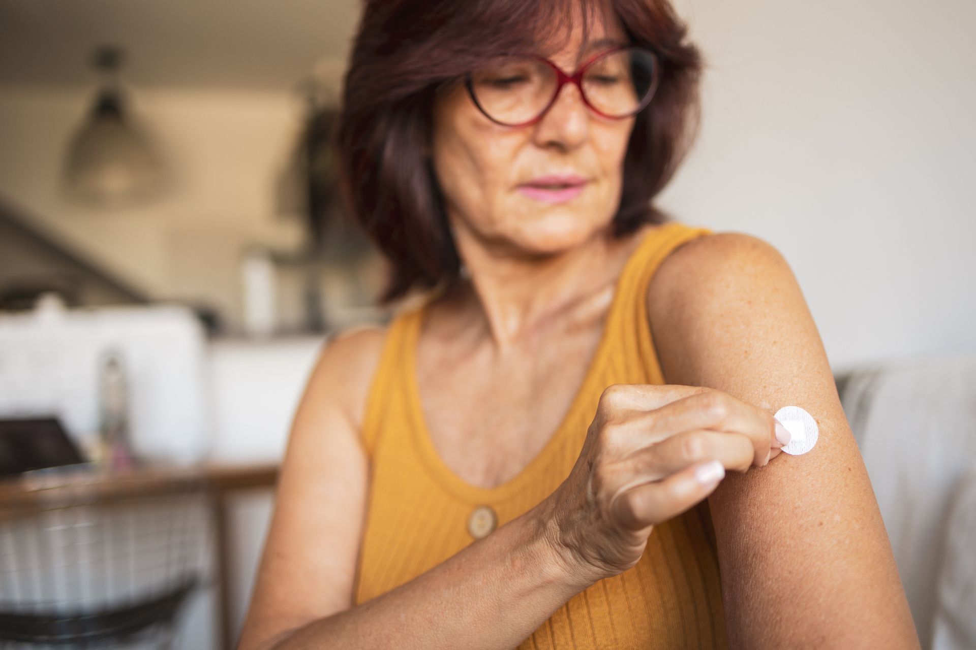 Woman applying a patch to her arm, indoors, wearing glasses and a yellow top.