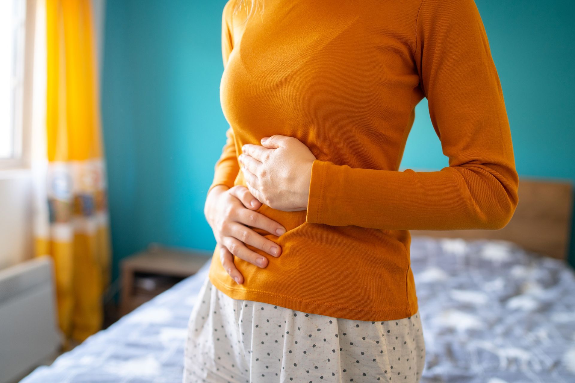 Woman holding stomach, possibly in pain. Indoors, wearing orange shirt and white pants, bedroom setting.