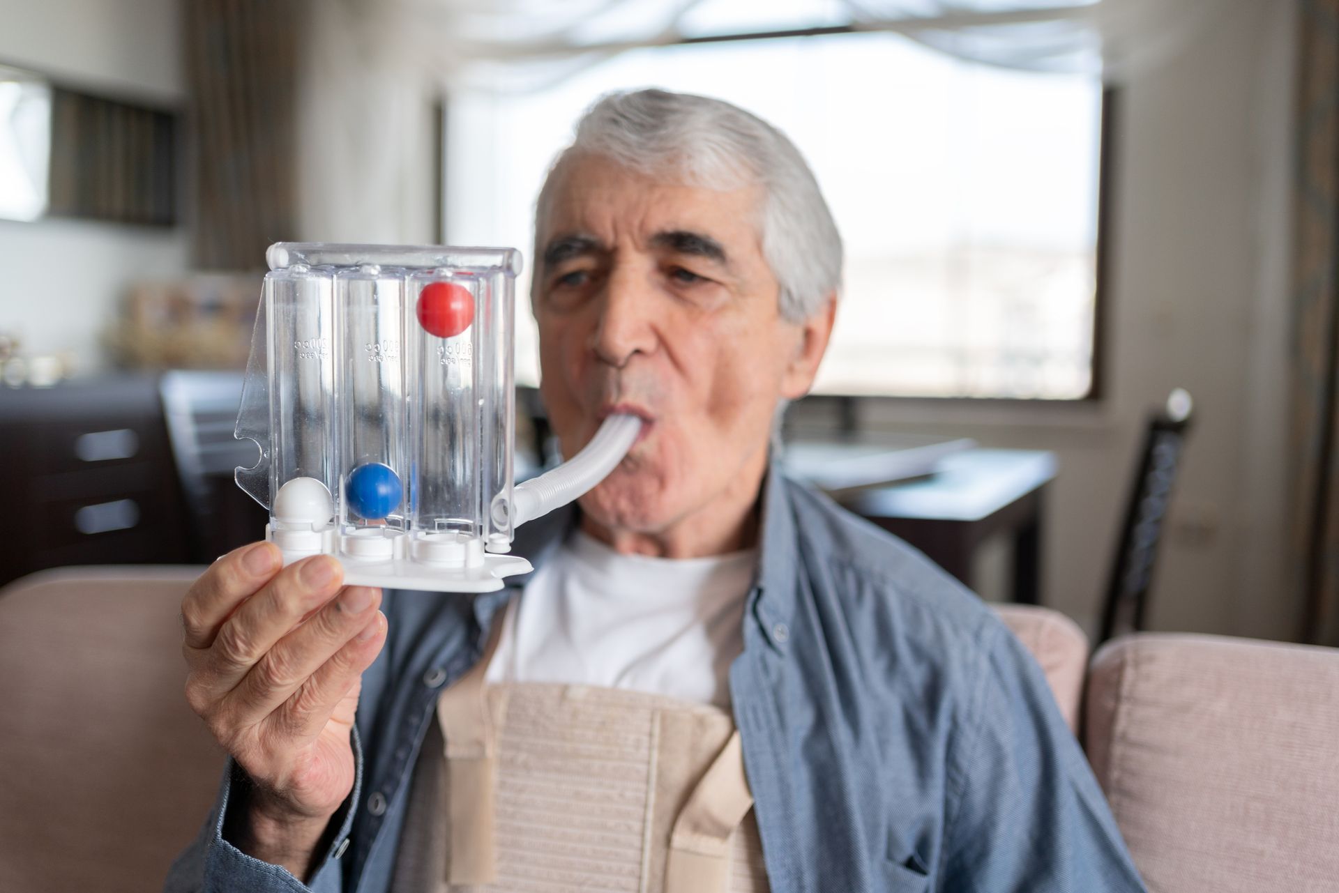 Older man using a breathing exerciser with red and blue balls indoors.