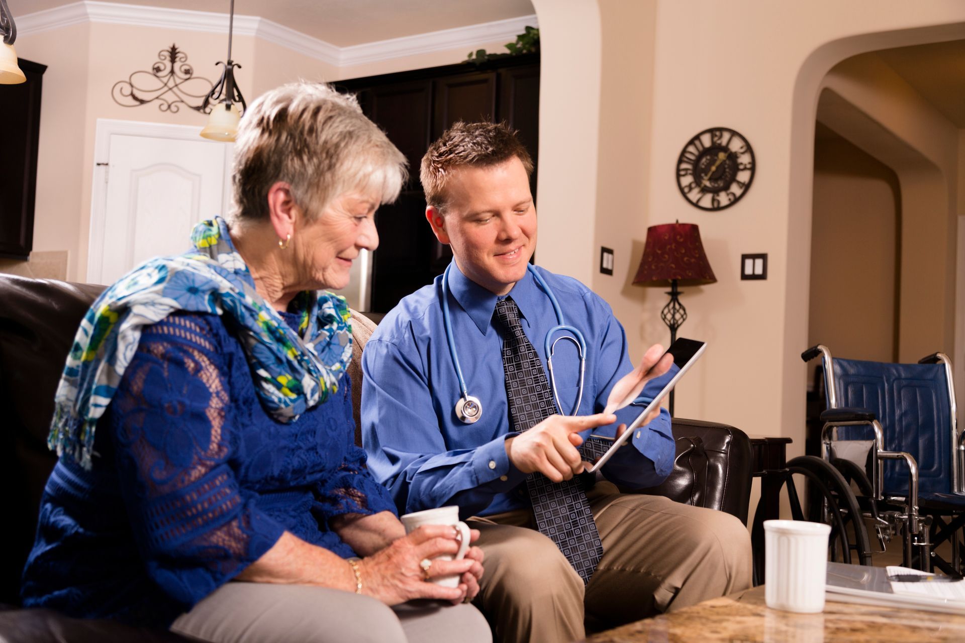 A doctor shows an elderly woman a tablet; they sit in a living room.