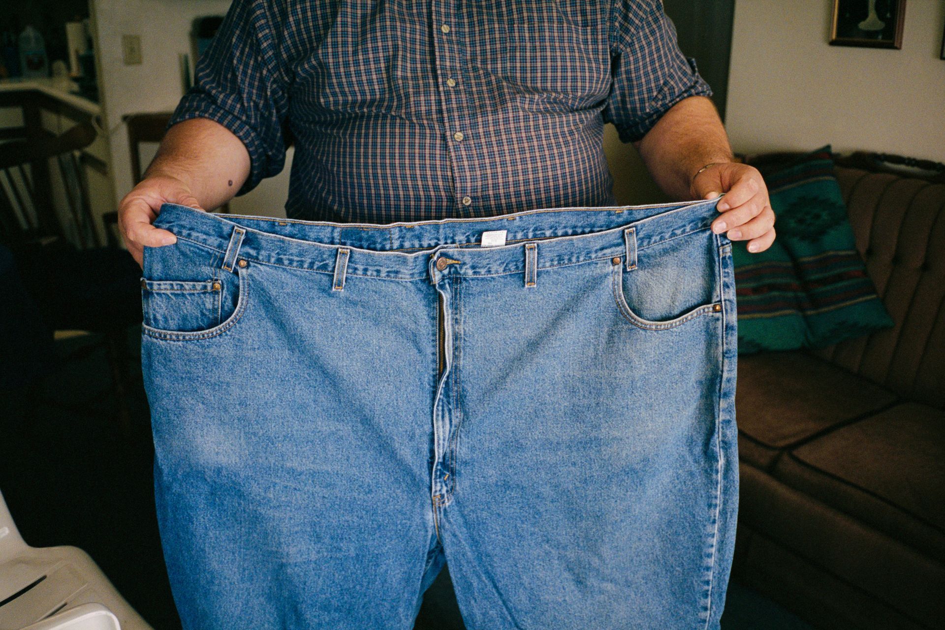 A man holds in front of him a pair of jeans that are of a much larger size than the one he is.